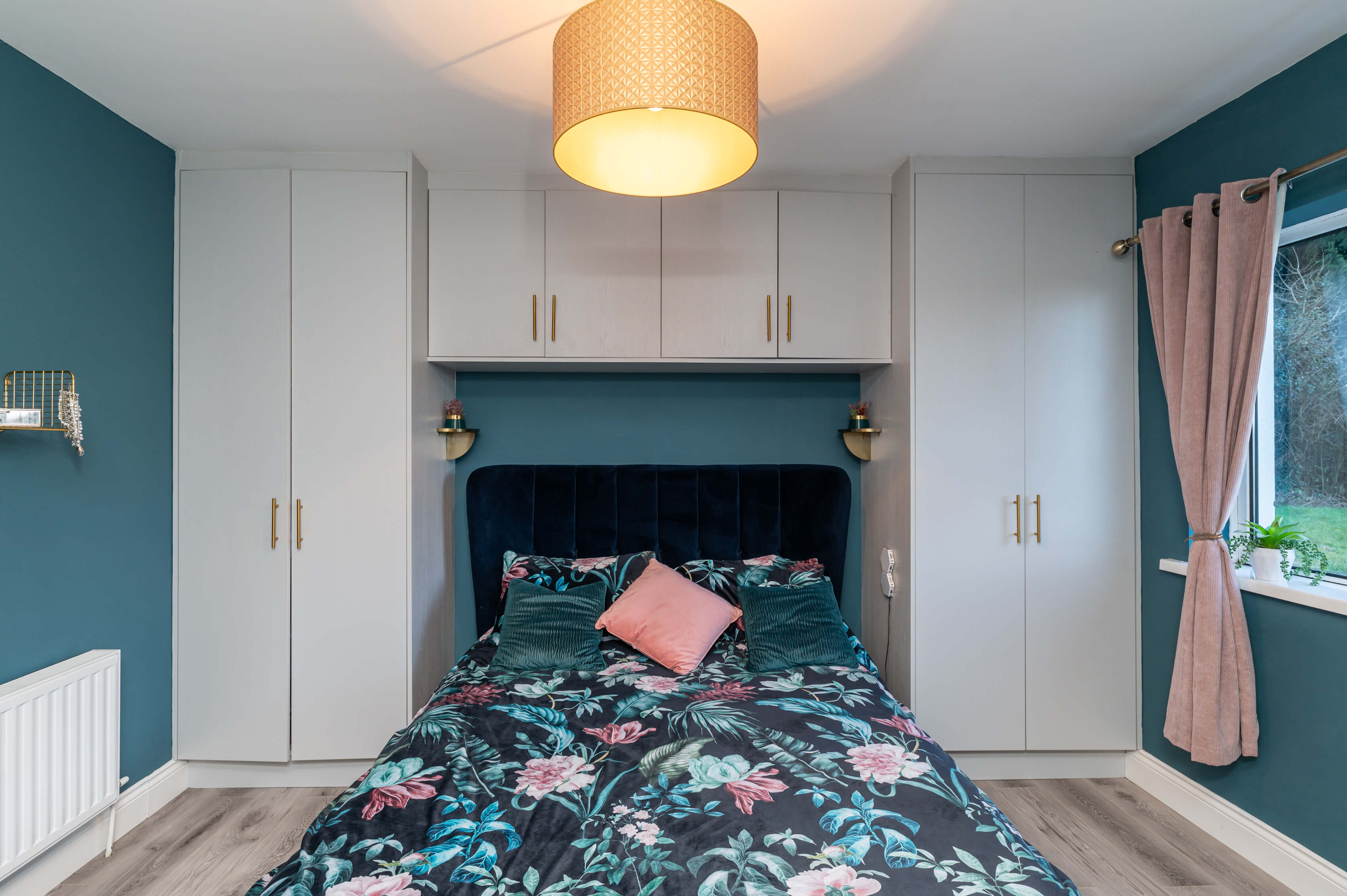 a master bedroom with teal green walls, a queen size bed with navy blue velevet frame and headboard. The wardrobes are grey with gold handles. There are 2 gold shelves either side of the bed. The curtians are a ribbed blush pink and the floor is grey laminate.